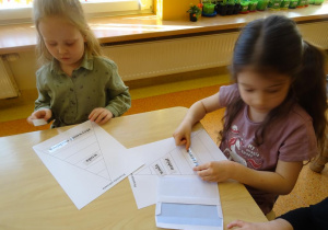 Dwie dziewczynki układają piramidy zdrowia z wykorzystaniem szablonów i napisami nazw produktów spożywczych.